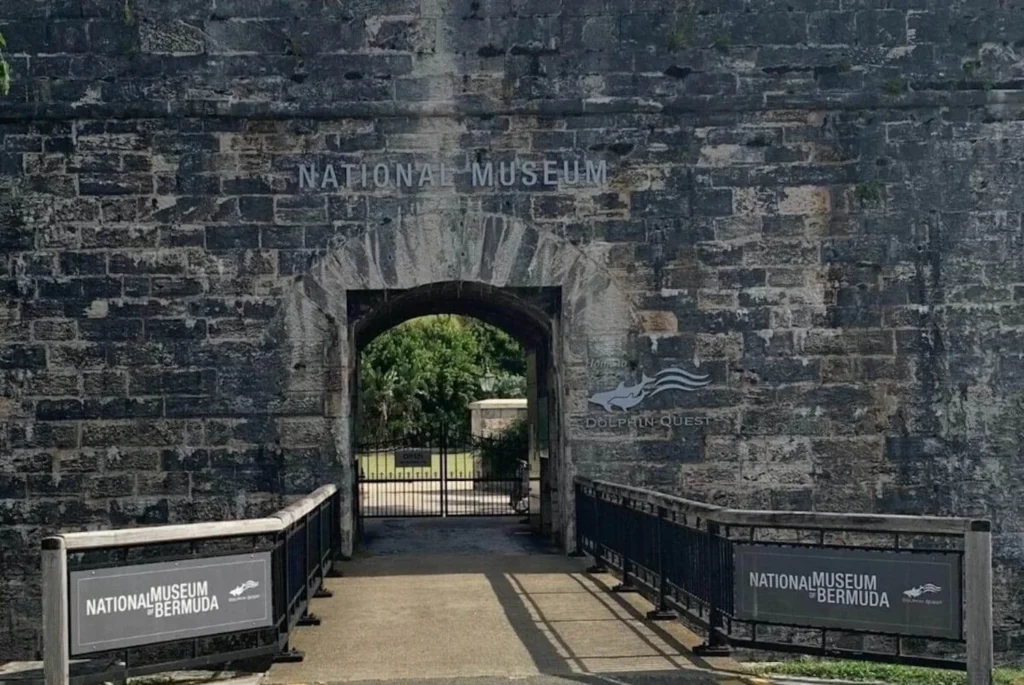 Stone entrance to the National Museum of Bermuda with signage for 'Dolphin Quest,' set against a clear day, showcasing the museum's historic architecture and cultural attractions.
