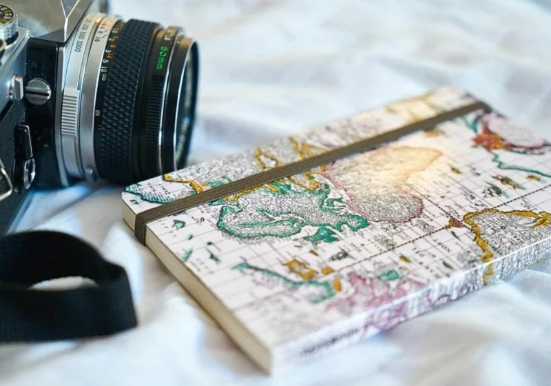 Close-up of a vintage camera with a detailed travel journal featuring a world map on its cover, resting on a soft white fabric surface.