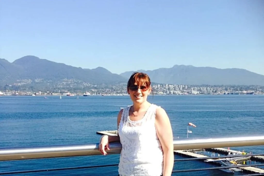 A smiling woman, Gemma Lawrence, in a white lace top leans on a railing with a stunning backdrop of the calm waters of Vancouver Harbor and the North Vancouver skyline under a clear blue sky, highlighting the beauty of the city's waterfront and mountainous landscape.