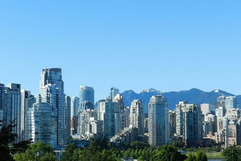 Skyline of Vancouver, Canada, with a mix of modern high-rises against a backdrop of snow-capped mountains and clear blue skies, showcasing the city's blend of urban development and natural beauty.