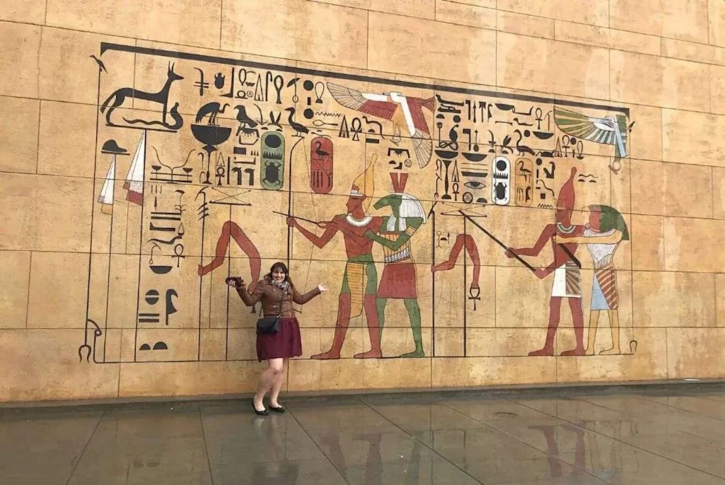 A woman, Gemma Lawrence, in a brown jacket and maroon skirt joyfully poses in front of a large wall mural depicting ancient Egyptian hieroglyphics and figures, likely at the Grauman's Egyptian Theatre in Hollywood.