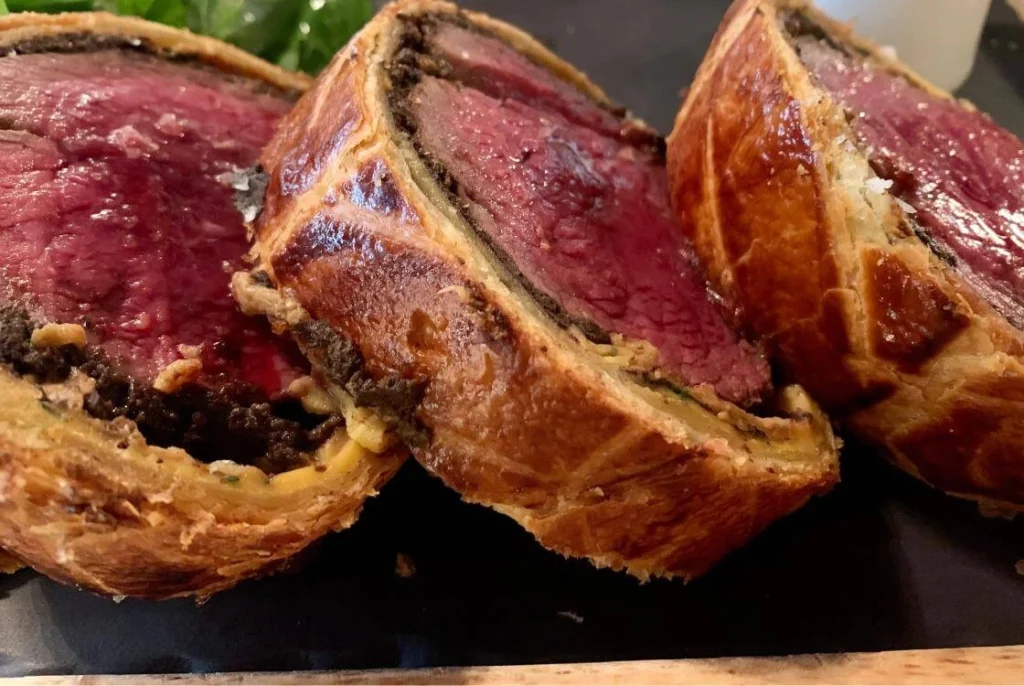 Sliced beef Wellington with golden puff pastry and pink tender beef filling served on a black board.