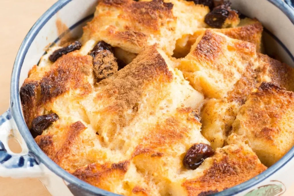 A warm, golden-brown bread and butter pudding in a ceramic dish, studded with dark raisins and dusted with cinnamon, ready to be served.