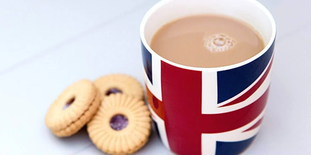 A cup of British tea with milk served in a Union Jack mug beside a few classic British biscuits on a white table, symbolizing traditional British tea culture.