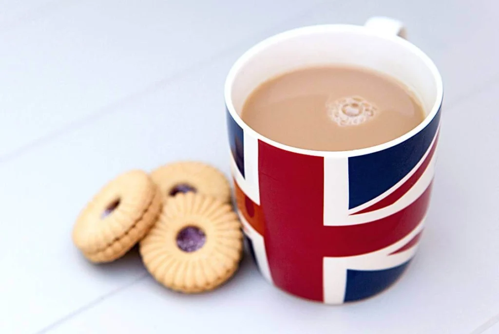 A cup of British tea with milk served in a Union Jack mug beside a few classic British biscuits on a white table, symbolizing traditional British tea culture.