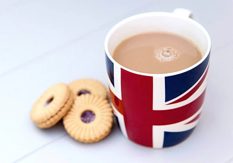 A cup of British tea with milk served in a Union Jack mug beside a few classic British biscuits on a white table, symbolizing traditional British tea culture.