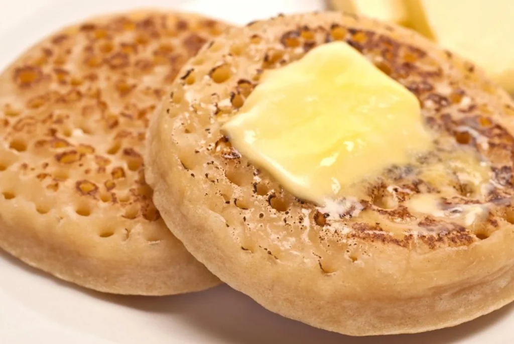 Two toasted crumpets with honeycomb-like interiors, the top one generously topped with melting butter, emphasizing the warm, comforting nature of this traditional British teatime treat.