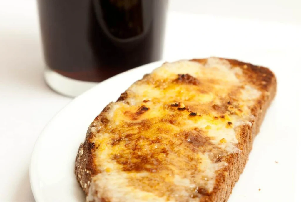 Toasted slice of bread topped with melted cheese served with a pint of dark ale in the background. 