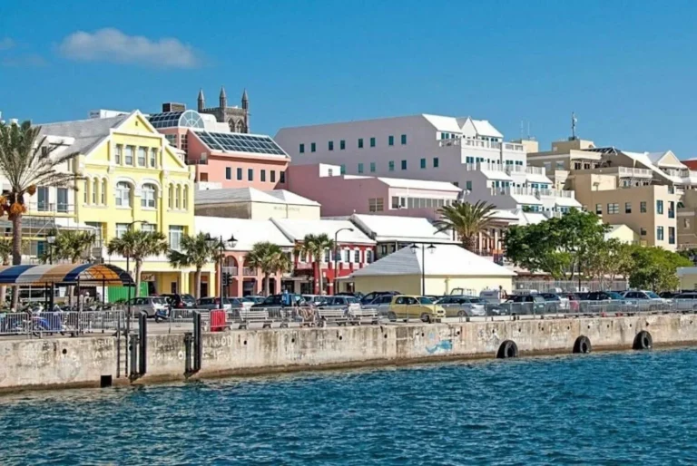 Colorful waterfront buildings along the harbor in the City of Hamilton, Bermuda, with clear blue waters in the foreground and a sunny sky above.