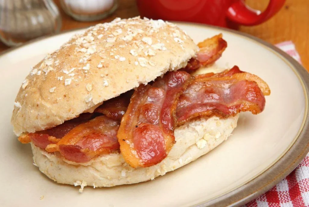 Classic British bacon roll on an oat-topped bun served on a plate beside a red teapot.