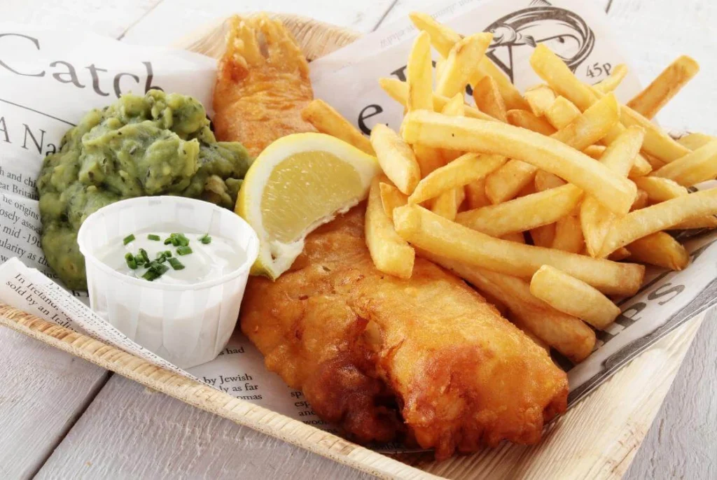 Traditional British fish and chips with mushy peas, tartar sauce, and lemon wedge served on newspaper-style paper.
