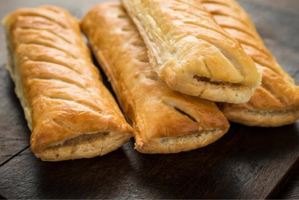 Freshly baked British sausage rolls with flaky golden pastry on a dark wooden board. 