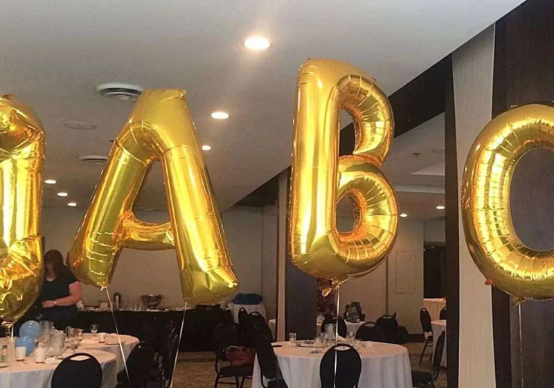 Golden helium balloon letters "IABC" floating in a conference room with set tables, suggesting an event or celebration related to the International Association of Business Communicators.