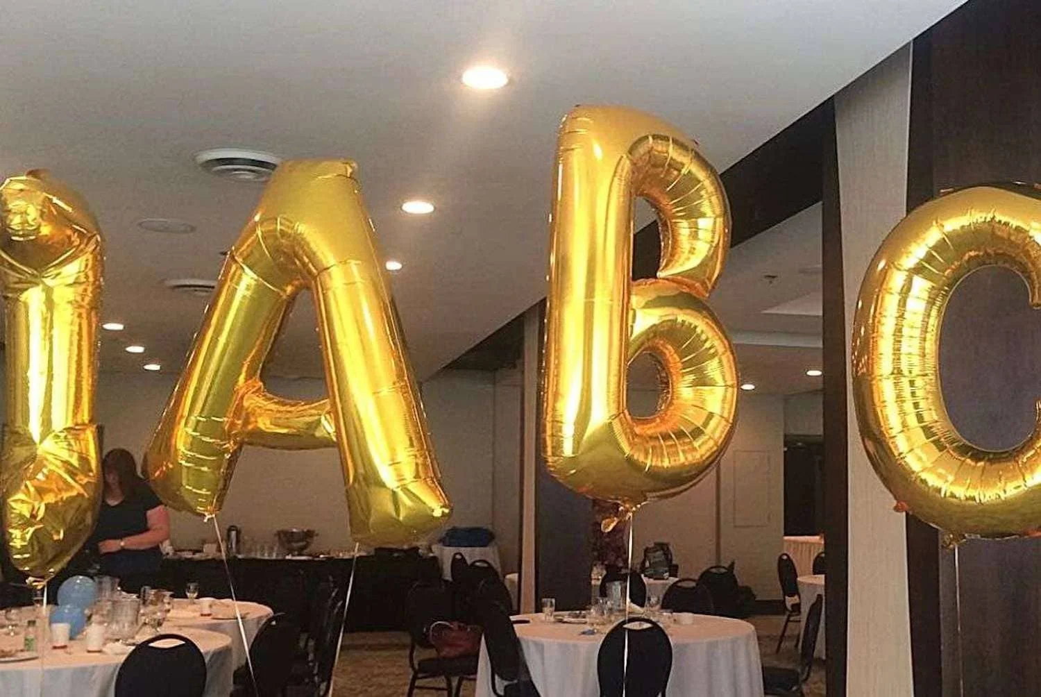 Golden helium balloon letters "IABC" floating in a conference room with set tables, suggesting an event or celebration related to the International Association of Business Communicators.
