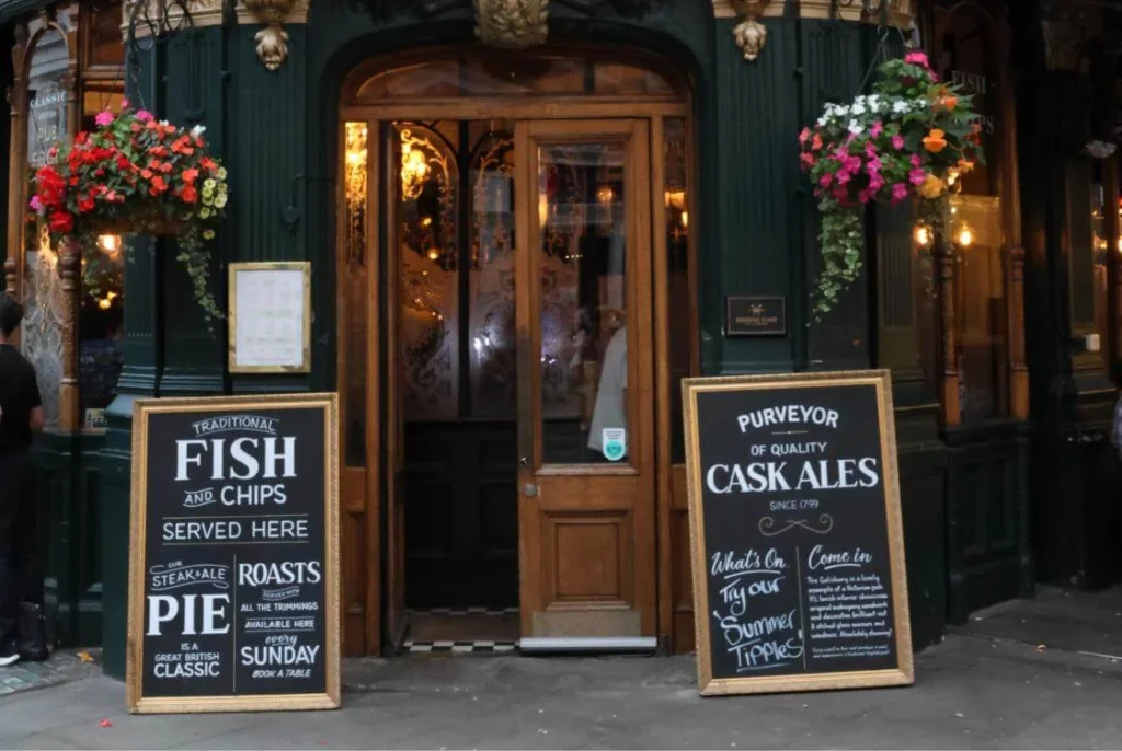 Classic London pub exterior with hanging flower baskets and chalkboard signs advertising fish and chips and cask ales. 