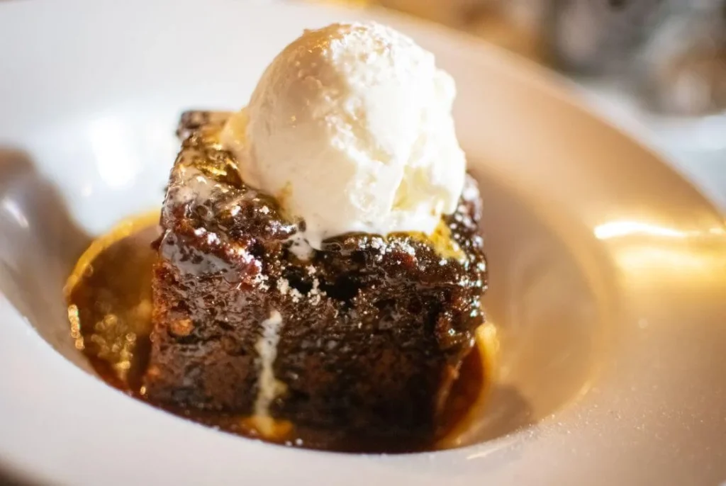 Classic British sticky toffee pudding topped with vanilla ice cream and drizzled with warm toffee sauce served in a white bowl.