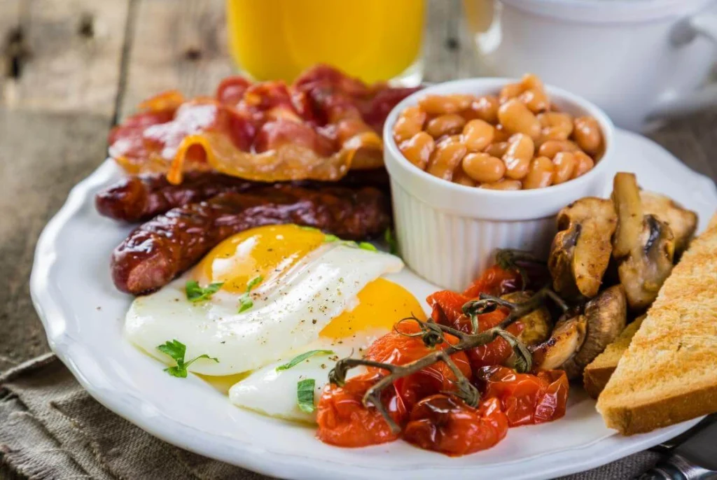 Traditional full English breakfast with fried eggs, bacon, sausages, baked beans, tomatoes, mushrooms, toast, and orange juice on a rustic wooden table. 