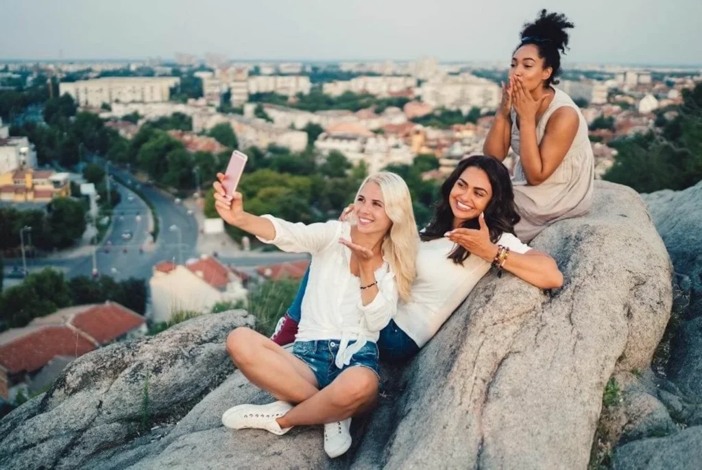 Three friends posing for a selfie on a rocky outcrop with a blurred cityscape in the background. One is holding the phone, another is making a peace sign, and the third is covering her mouth with her hands in a gesture of excitement.
