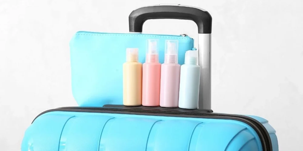 A light blue hard-shell carry-on suitcase with a matching toiletry bag and an assortment of pastel-colored travel-sized bottles for personal care products on top, against a neutral background.
