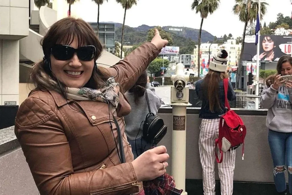 A woman, Gemma Lawrence, in a brown leather jacket and sunglasses smiles while pointing towards the Hollywood sign in the distance, surrounded by other tourists and a viewfinder on a bustling observation deck.