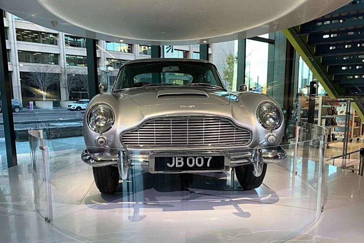 A classic silver Aston Martin, often associated with the James Bond franchise, is on display with the license plate "JB 007", indicating its iconic status as a spy vehicle, showcased in a modern indoor setting at the Spy Museum in Washington DC.