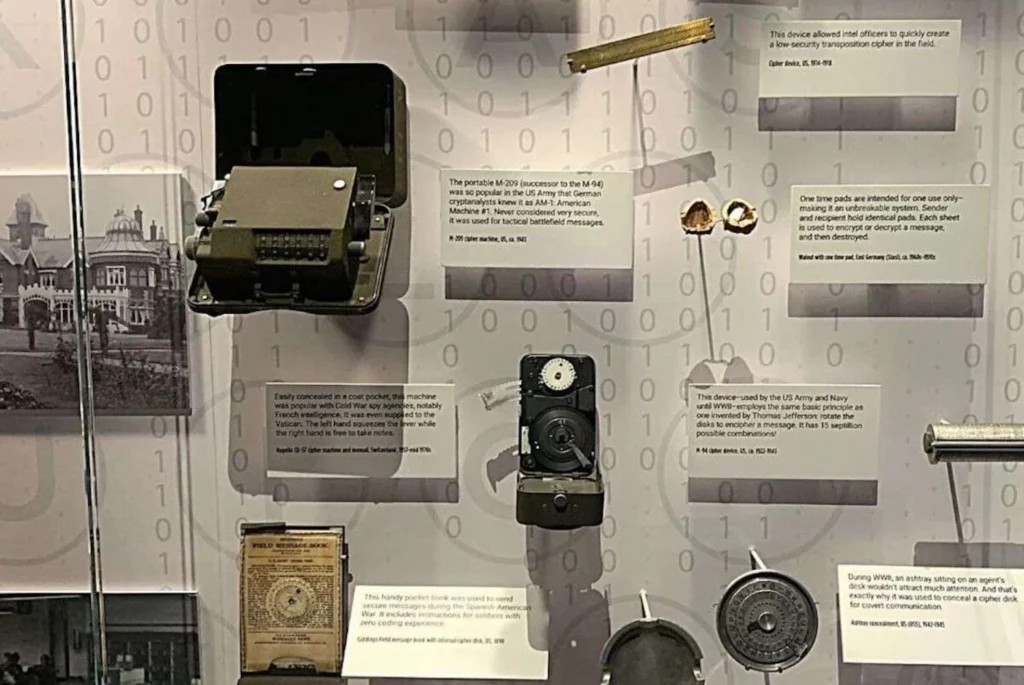A display of espionage devices, with a central focus on a portable M-209 cipher machine from World War II, known for its use by the US Army and other agencies. Various cryptographic tools and gadgets are showcased, each with explanatory texts, including cipher disks and one-time pad sheets. The background features binary code graphics, adding to the secretive and technical ambiance of the exhibit at the Spy Museum in Washington DC.