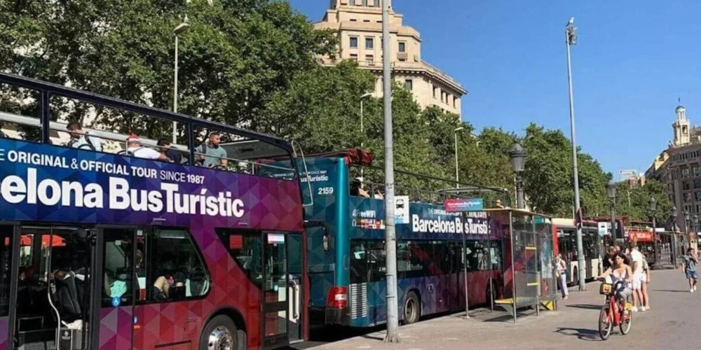 A vibrant street scene featuring the 'Barcelona Bus Turistic', the city's official hop-on hop-off tour service, with passengers on the open-top deck enjoying the sights, flanked by pedestrians and cyclists on a sunny day.