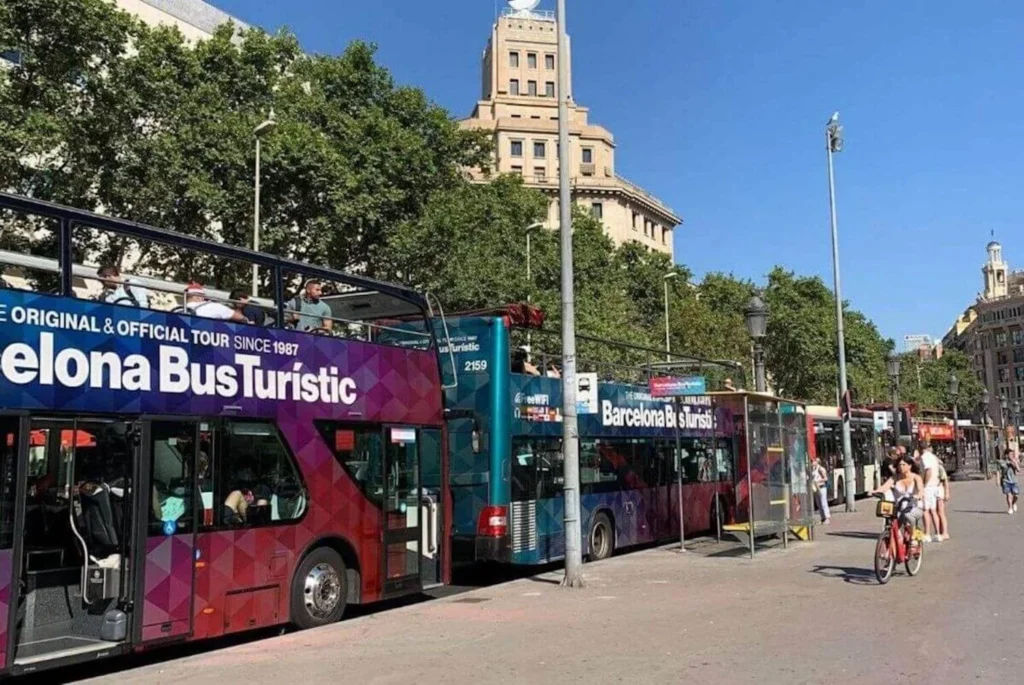 A vibrant street scene featuring the 'Barcelona Bus Turistic', the city's official hop-on hop-off tour service, with passengers on the open-top deck enjoying the sights, flanked by pedestrians and cyclists on a sunny day.