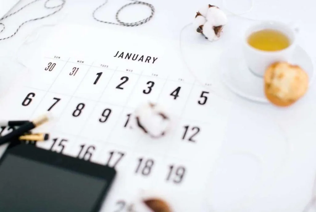 A January calendar page in soft focus with planning accessories, including a white teacup, a cotton plant, and a stylus on a digital tablet, set against a white background for a clean and organized new year planning theme.