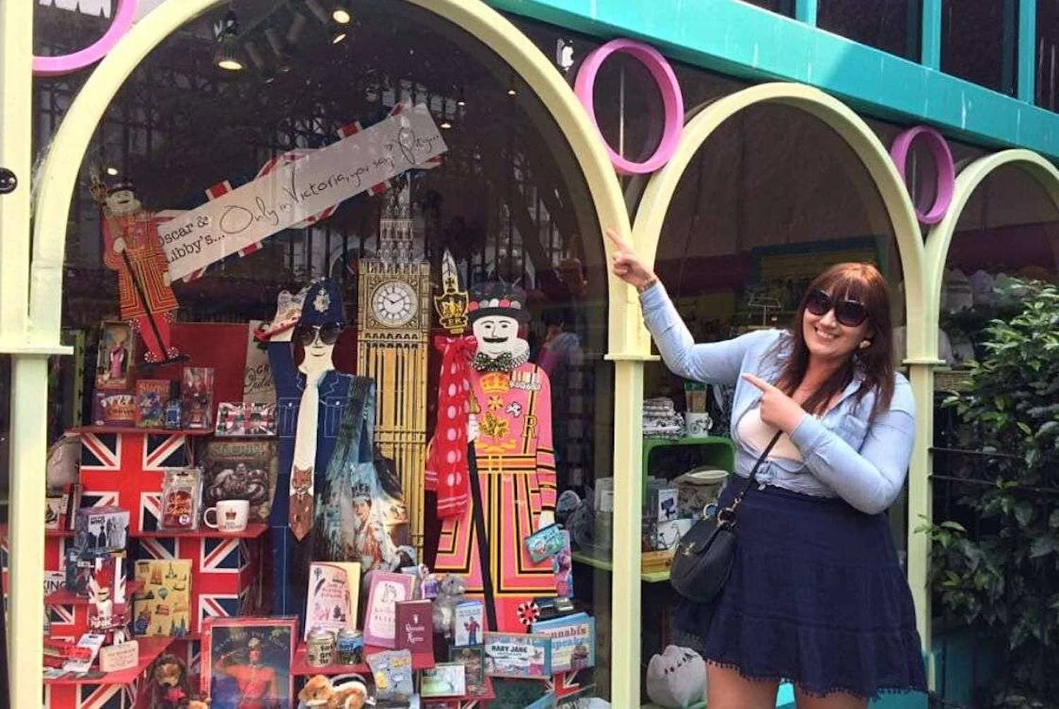 A woman, Gemma Lawrence with sunglasses stands smiling and pointing at a colorful storefront in Victoria, BC that's adorned with British cultural icons, including a Beefeater and a cutout of a British Royal Guard, suggesting a shop specializing in UK-themed goods.
