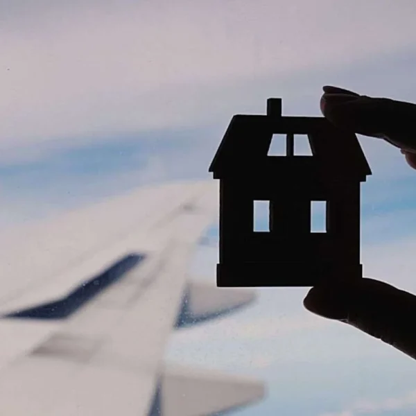 A silhouette of a hand holding a small house-shaped object against an airplane window, with the blurred wing in the background, evoking a sense of homesickness or longing for home while traveling.