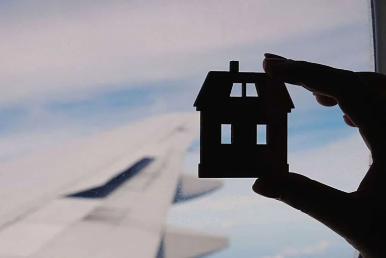 A silhouette of a hand holding a small house-shaped object against an airplane window, with the blurred wing in the background, evoking a sense of homesickness or longing for home while traveling.