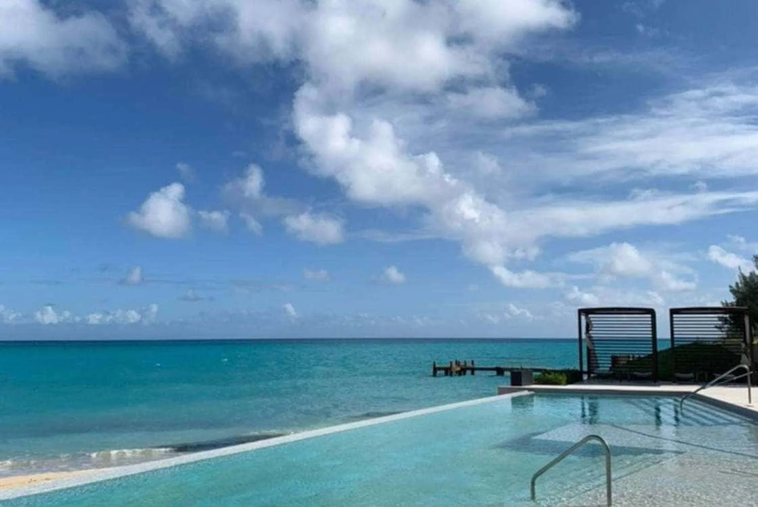 An infinity pool at the St Regis in Bermuda blends seamlessly into the serene turquoise sea, with a clear blue sky dotted with fluffy clouds above, creating a tranquil and luxurious beachside escape.