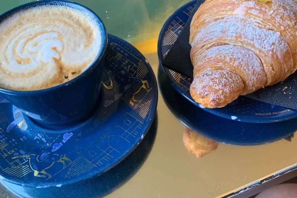 A delightful breakfast scene featuring a freshly brewed cup of cappuccino with a beautiful foam swirl on top, accompanied by a flaky, golden-brown croissant dusted with powdered sugar. Both items are served on a deep blue saucer and plate set, with intricate gold patterns that add a touch of elegance to the morning fare. The reflection on the table suggests a sunny ambience, possibly in an outdoor setting, inviting a peaceful start to the day.