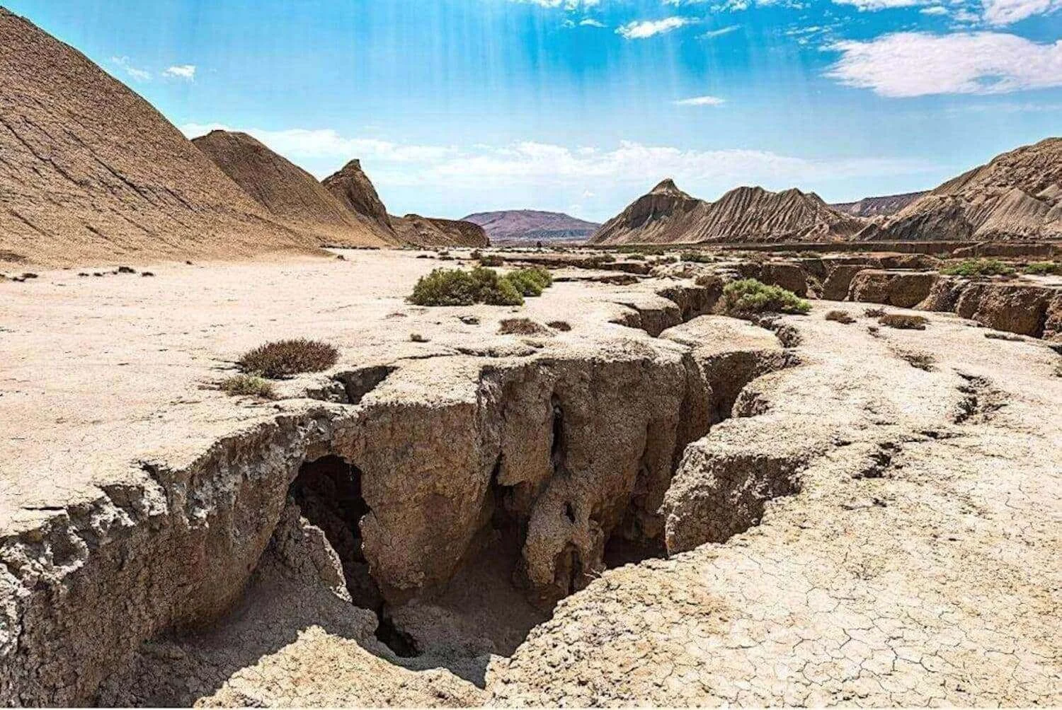 A stark desert landscape characterized by deep crevices and fissures that might suggest geological activity such as an earthquake. The arid environment is dotted with sparse vegetation, and dramatic sedimentary rock formations rise in the background under a bright blue sky. Sunlight streams down, highlighting the textured ground and the vastness of the open space, creating a scene of natural drama and the raw beauty of an untamed wilderness.