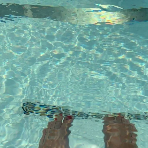 A person's feet submerged in the clear, tranquil waters of a swimming pool at the St Regis in Bermuda. The water's surface is calm, with sunlight casting shimmering patterns onto the pool floor, creating a peaceful and relaxing atmosphere. It's a moment captured during leisure time, evoking the pleasure of a refreshing dip on a sunny day.
