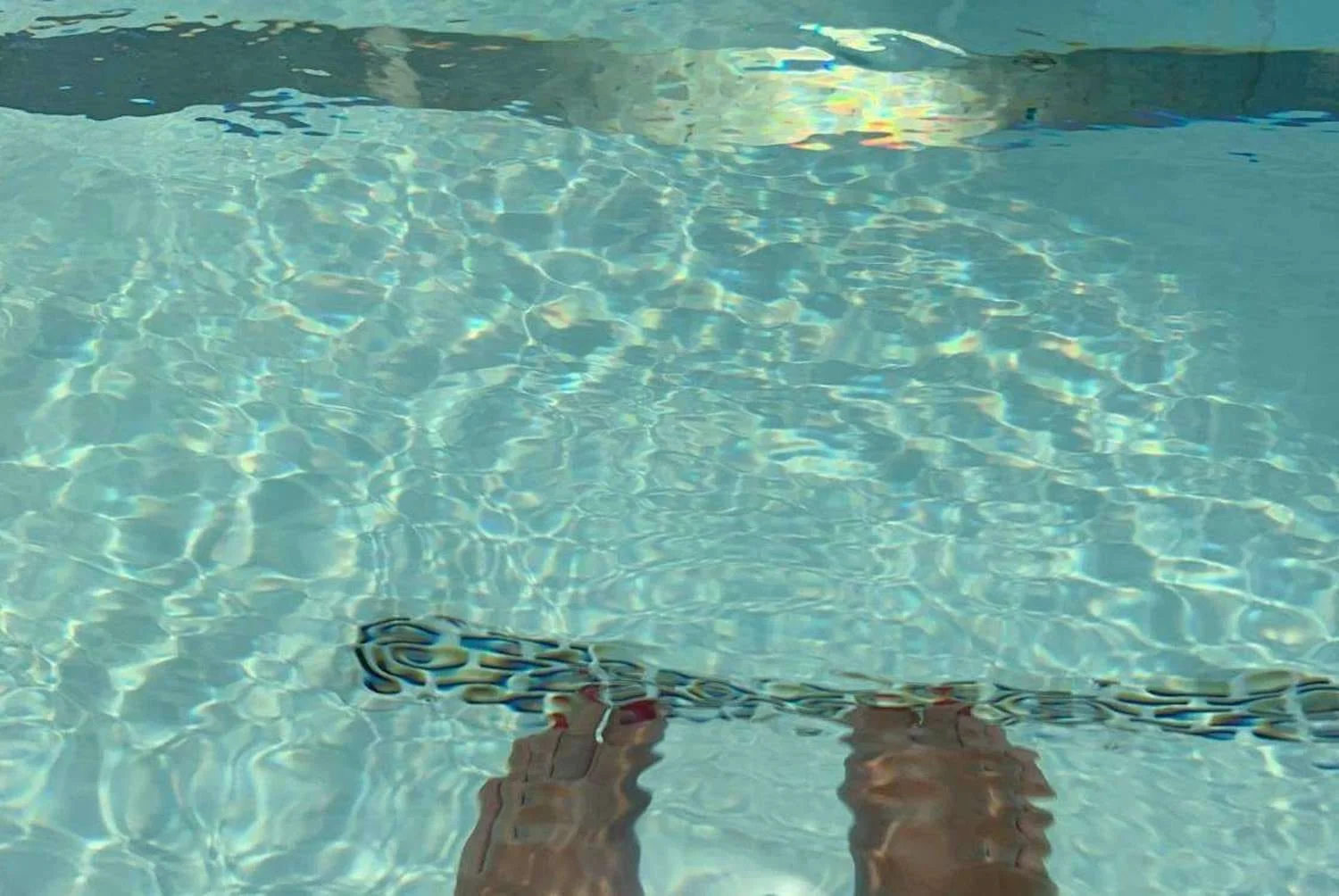 A person's feet submerged in the clear, tranquil waters of a swimming pool at the St Regis in Bermuda. The water's surface is calm, with sunlight casting shimmering patterns onto the pool floor, creating a peaceful and relaxing atmosphere. It's a moment captured during leisure time, evoking the pleasure of a refreshing dip on a sunny day.