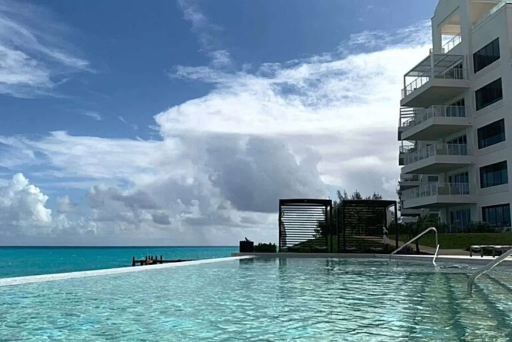 A luxurious hotel infinity pool at the St Regis in Bermuda overlooking the serene blue waters of the ocean. The pool's edge seamlessly blends with the sea, creating an endless water landscape. Fluffy white clouds adorn the sky, complementing the tranquil scene. A modern hotel building rises on the right, offering guests a spectacular view from each balcony. This setting suggests a peaceful retreat in a tropical location where one can relax and take in the natural beauty.