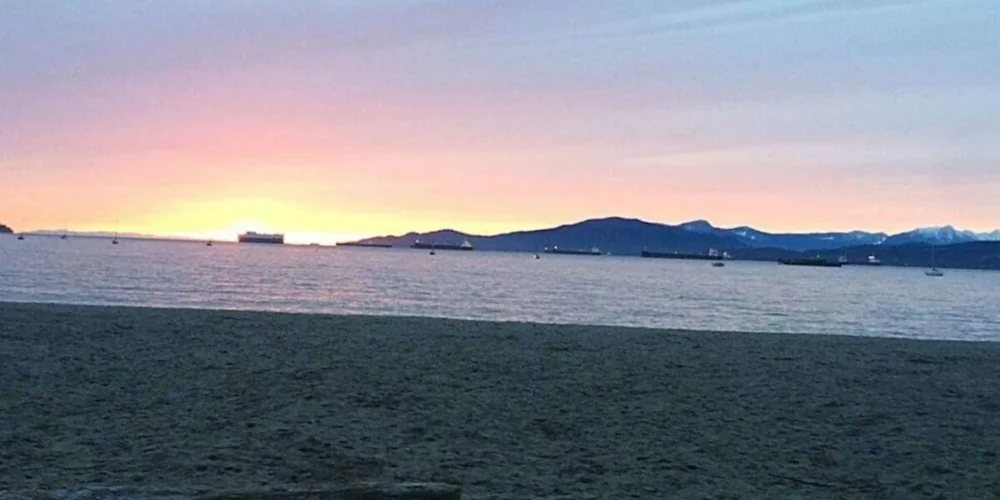 A tranquil beach scene at sunset in Vancouver, Canada. The sky is painted in soft hues of pink and purple as the sun dips below the horizon, casting a warm glow that reflects off the calm sea. Several ships are visible in the distance, anchored or slowly moving across the water, silhouetted against the fading light. The beach is empty, suggesting a peaceful end to the day, and the presence of a large driftwood log adds a natural element to the foreground, inviting viewers to imagine themselves sitting and watching the serene sunset. The scene evokes a sense of calm and the beauty of nature's displays.