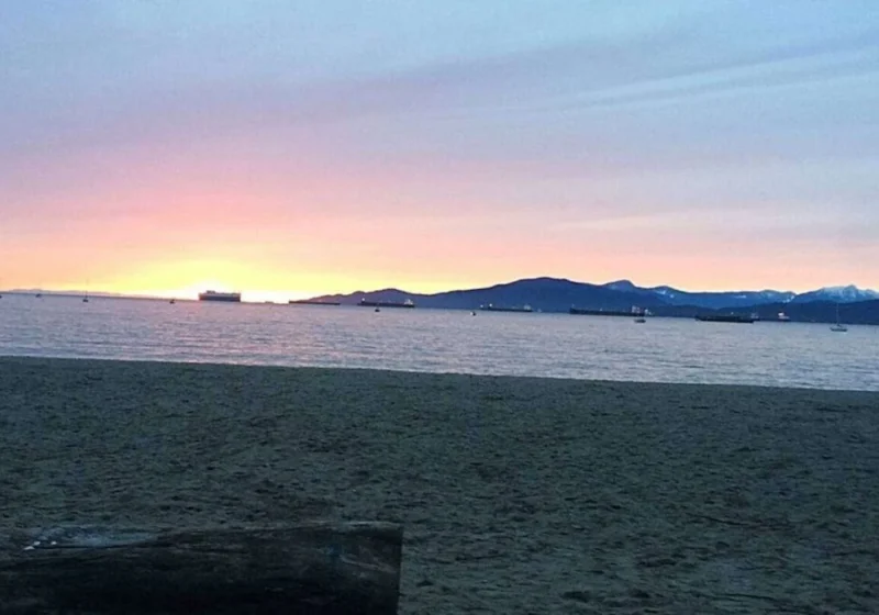 A tranquil beach scene at sunset in Vancouver, Canada. The sky is painted in soft hues of pink and purple as the sun dips below the horizon, casting a warm glow that reflects off the calm sea. Several ships are visible in the distance, anchored or slowly moving across the water, silhouetted against the fading light. The beach is empty, suggesting a peaceful end to the day, and the presence of a large driftwood log adds a natural element to the foreground, inviting viewers to imagine themselves sitting and watching the serene sunset. The scene evokes a sense of calm and the beauty of nature's displays.