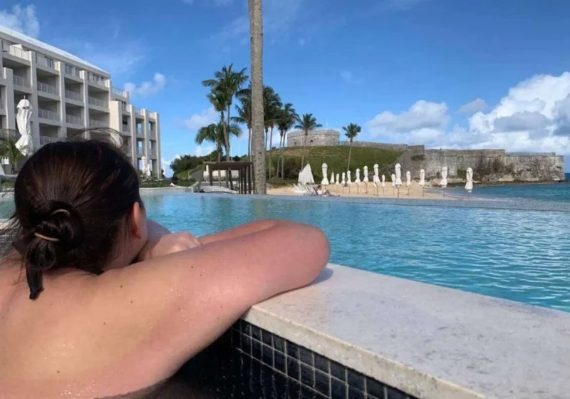 A woman, Gemma Lawrence, enjoying a serene poolside view at a luxurious resort in Bermuda. The infinity pool leads the eye towards an impressive coastal fortification under a bright blue sky scattered with clouds. The pool area is lined with neatly arranged sun loungers under closed white umbrellas, suggesting an atmosphere of relaxation and tranquility. Palm trees rise above the scene, adding a tropical vibe to this picturesque setting. It's a snapshot of a perfect holiday moment, blending historical architecture with modern leisure.
