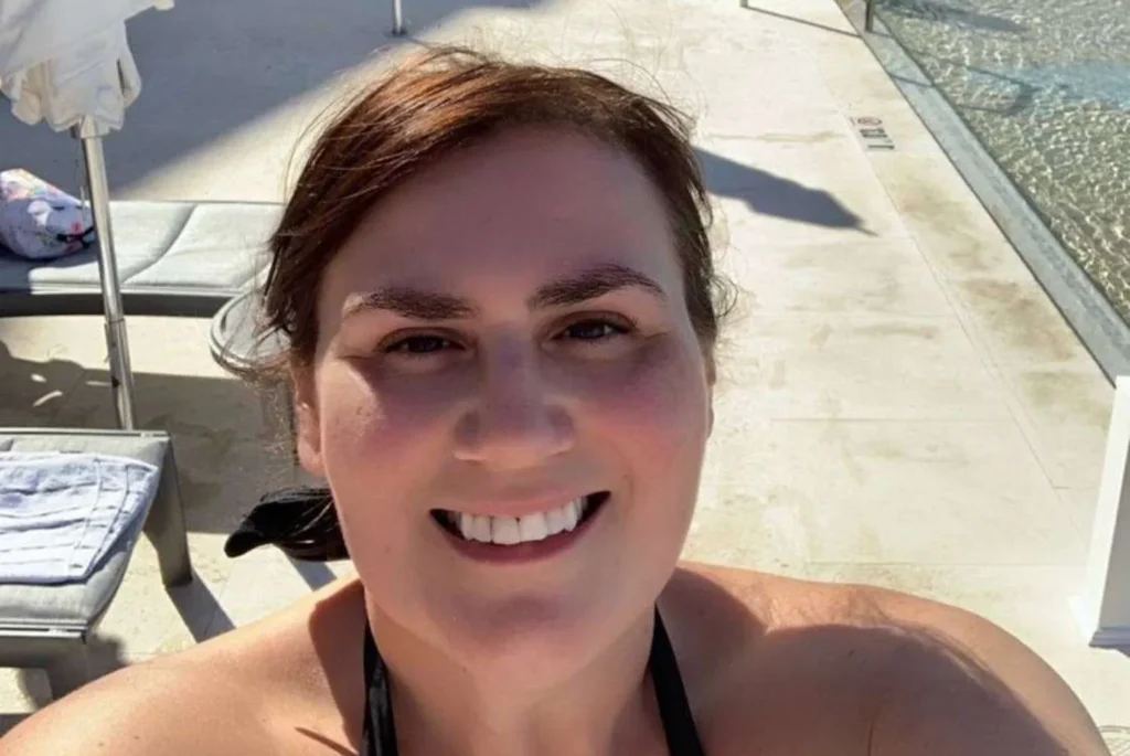 A joyful selfie of a woman, Gemma Lawrence, relaxing by the pool. She is smiling warmly at the camera with a bright, sunlit backdrop highlighting her features. The poolside setting is suggested by the glimpse of water and sun loungers in the background, indicating a leisurely day spent enjoying the outdoors. It's a personal and happy moment, likely from a vacation or a day off, where the person is soaking up the sunshine and the laid-back atmosphere.