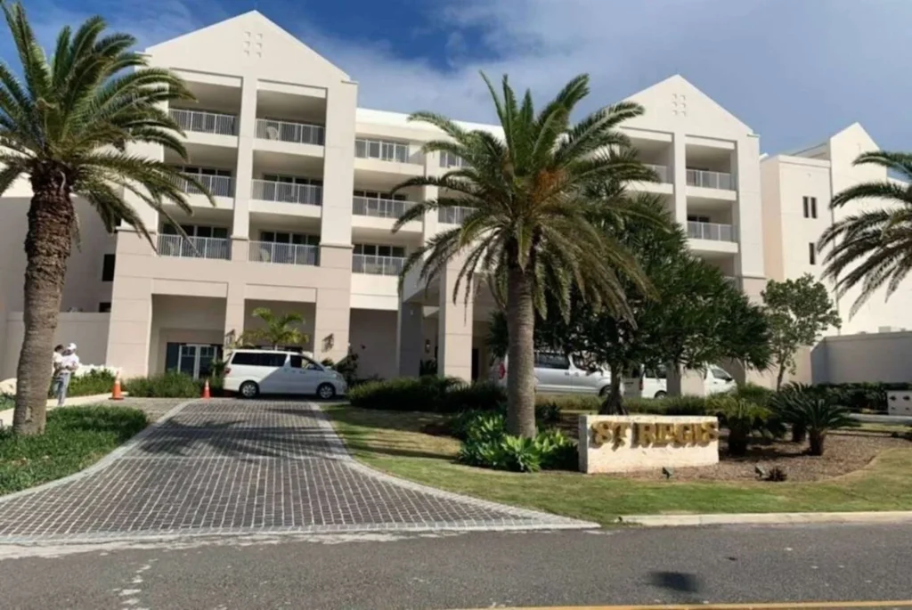 The entrance to the St. Regis hotel, marked by a prominently displayed sign amidst a landscaped area with palm trees. The architecture of the building, visible in the background, exhibits a modern design with neutral tones and multiple balconies. The clear sky suggests a pleasant day, and the presence of a parked car hints at the accessibility and hospitality awaiting guests at this luxurious destination in Bermuda.