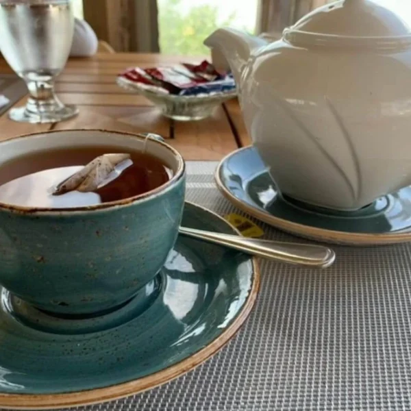 A tranquil tea time scene, showcasing an earthy-toned ceramic cup of tea with a matching saucer, complemented by a sleek white teapot resting on a similar hued saucer. The tea bag's tag peeks over the cup's edge, suggesting a freshly steeped warm brew. In the background, there's a hint of a laid-back dining setting with soft natural light filtering through a window, enhancing the serene ambiance perfect for a leisurely afternoon break.
