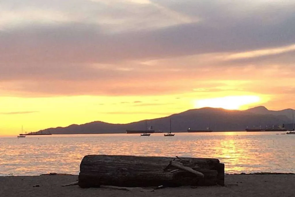 A serene sunset at a Vancouver beach, with warm hues of orange and yellow painting the sky. Silhouetted mountains form a tranquil backdrop, while the calm sea reflects the sunset’s glow. A few boats are anchored in the distance, contributing to the peaceful scene. A large driftwood log rests on the sandy shore in the foreground, inviting viewers to imagine sitting and soaking in the picturesque moment.