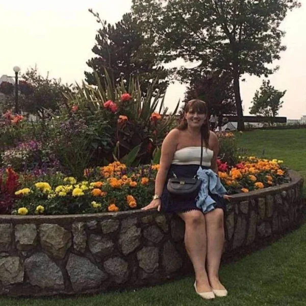The photo shows a woman, Gemma Lawrence, posing with a cheerful smile in a lush garden filled with a vibrant assortment of flowers. The stone-edged flowerbed boasts a colorful display of flora, indicative of the well-maintained public gardens found in Victoria, BC. In the background, the scene is lively with people enjoying the outdoor space, and there's a glimpse of the city's architecture, contributing to the urban park's charm. It's a casual, yet picturesque snapshot of a day spent among the natural beauty that Victoria is known for.