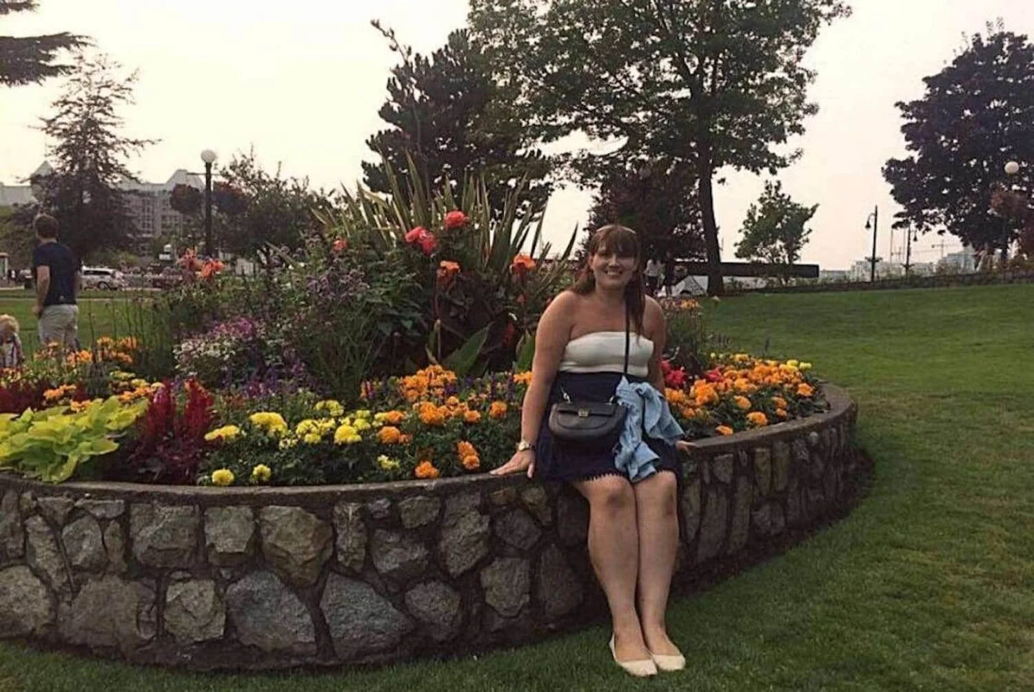 The photo shows a woman, Gemma Lawrence, posing with a cheerful smile in a lush garden filled with a vibrant assortment of flowers. The stone-edged flowerbed boasts a colorful display of flora, indicative of the well-maintained public gardens found in Victoria, BC. In the background, the scene is lively with people enjoying the outdoor space, and there's a glimpse of the city's architecture, contributing to the urban park's charm. It's a casual, yet picturesque snapshot of a day spent among the natural beauty that Victoria is known for.