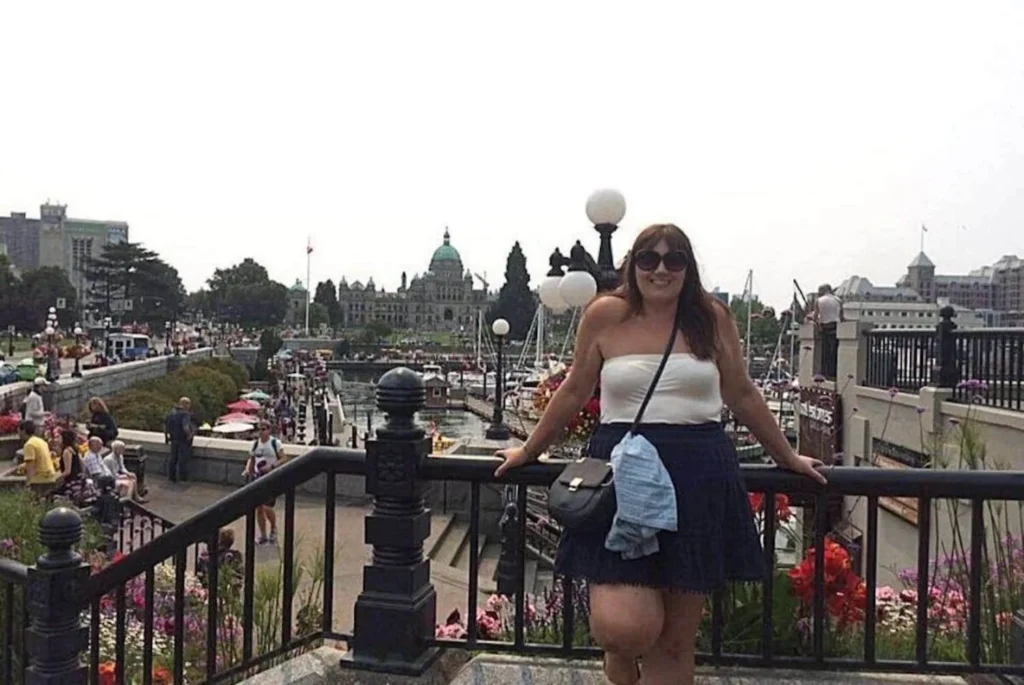 The image features a woman, Gemma Lawrence, smiling for the camera, standing at a viewpoint with a scenic backdrop. She is casually dressed for a day out, with sunglasses perched on her head, suggesting sunny weather. Behind her, the bustling waterfront walkway is lined with vibrant flower beds, and the iconic architecture of the British Columbia Parliament Buildings is visible in the distance, indicating this is likely in Victoria, BC. The scene captures a lively atmosphere with visitors enjoying the waterside promenade, contributing to the city's charm and vibrancy.