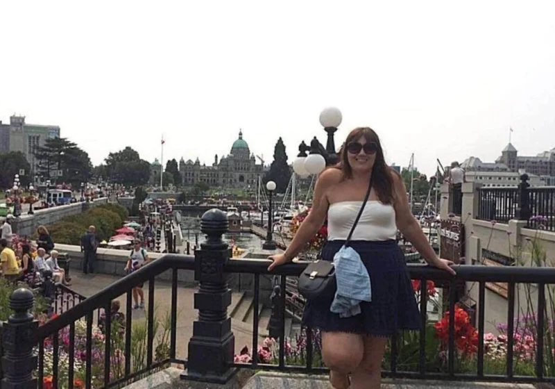 The image features a woman, Gemma Lawrence, smiling for the camera, standing at a viewpoint with a scenic backdrop. She is casually dressed for a day out, with sunglasses perched on her head, suggesting sunny weather. Behind her, the bustling waterfront walkway is lined with vibrant flower beds, and the iconic architecture of the British Columbia Parliament Buildings is visible in the distance, indicating this is likely in Victoria, BC. The scene captures a lively atmosphere with visitors enjoying the waterside promenade, contributing to the city's charm and vibrancy.