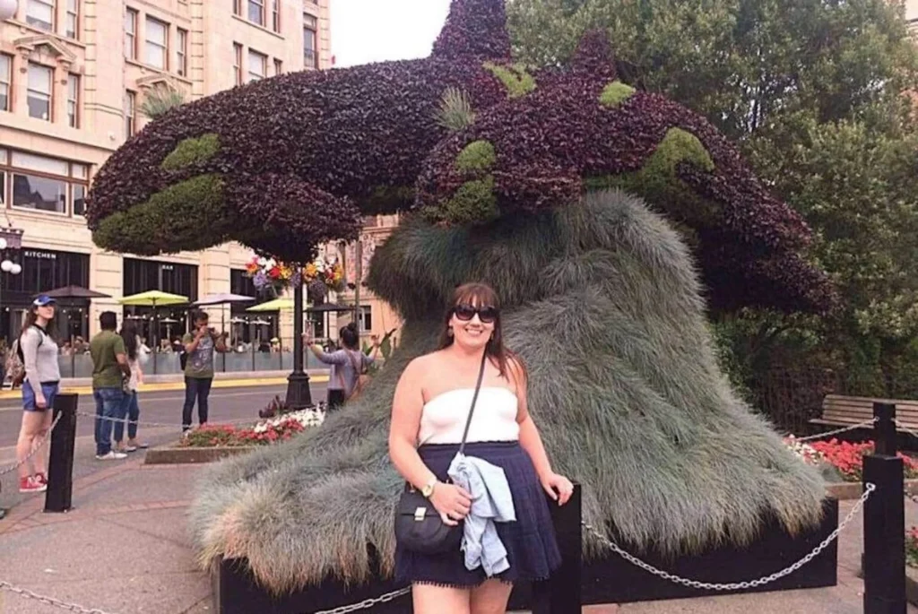 In the photograph, a woman, Gemma Lawrence poses with a smile in front of an imaginative topiary shaped like a whale, a creative blend of horticulture and artistry typical of Victoria, BC's charm. The whimsical greenery sculpture provides a unique backdrop, with the bustling city street life visible in the distance. This setting suggests a popular tourist spot, where visitors can enjoy the intersection of urban vibrancy and botanical whimsy. The image captures a moment of leisure and enjoyment in a vibrant, green urban space.